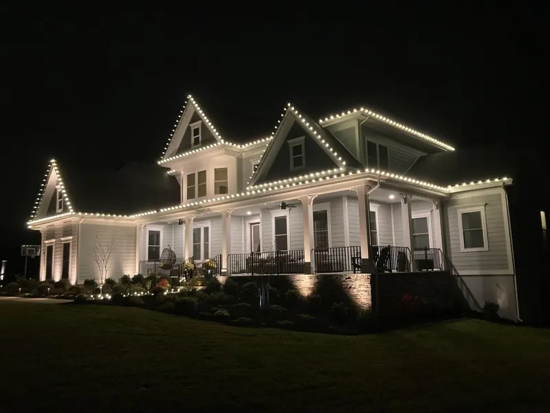 Large white farmhouse with roofline, porch and gables outlined in warm white Christmas lights at night