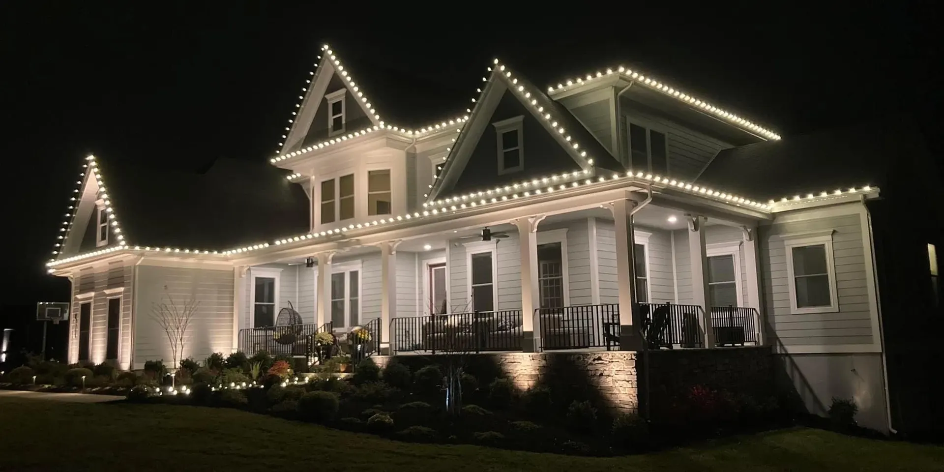 Large white farmhouse with its roofline and gables outlined in warm white Christmas lights at night